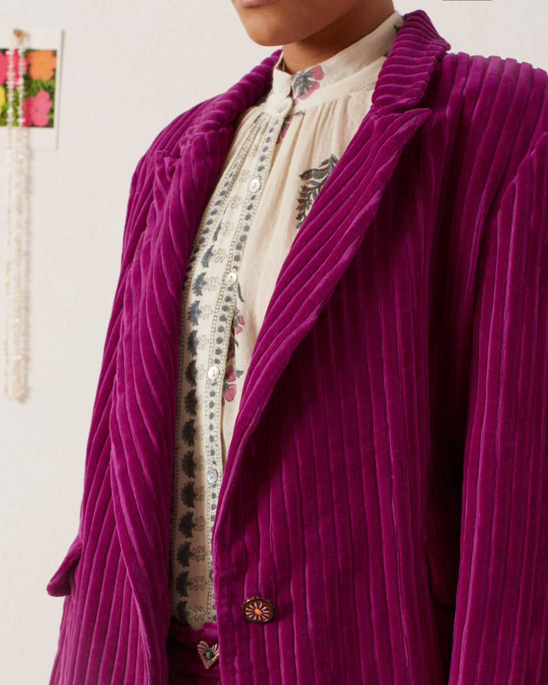 A person is wearing a Dree Violet Velvet Blazer over a cream blouse with a delicate floral and geometric design. Part of the room's decor, including colorful wall art, is visible in the background.