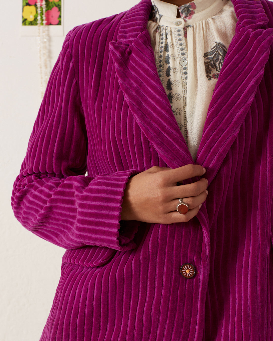 A person wearing the Dree Violet Velvet Blazer over a white blouse with ornate designs skillfully adjusts the blazer with one hand, revealing a ring on their finger. The background is a plain wall adorned with a small decorative item.