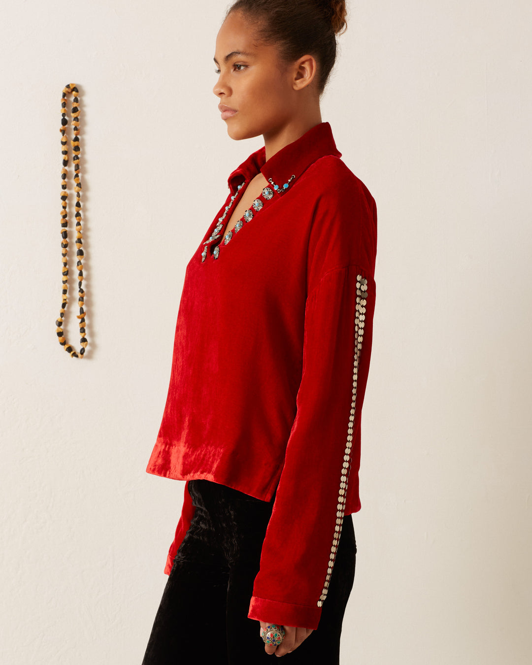 A person wearing the Dylan Ruby Velvet Tunic, adorned with decorative beads and embellishments, gazes to the side while standing against a plain white wall. A beaded necklace with Western-inspired flair is displayed on the wall in the background.