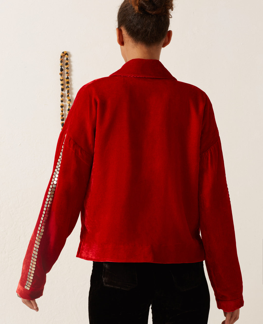 A person wearing the Dylan Ruby Velvet Tunic, styled with decorative trim on one sleeve, stands facing a white wall adorned with a hanging beaded accessory.