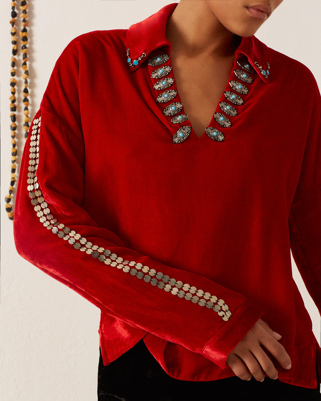 A person in a Western-inspired Dylan Ruby Velvet Tunic with decorative patterns on the collar and sleeves stands in front of a white wall adorned with beaded necklaces.