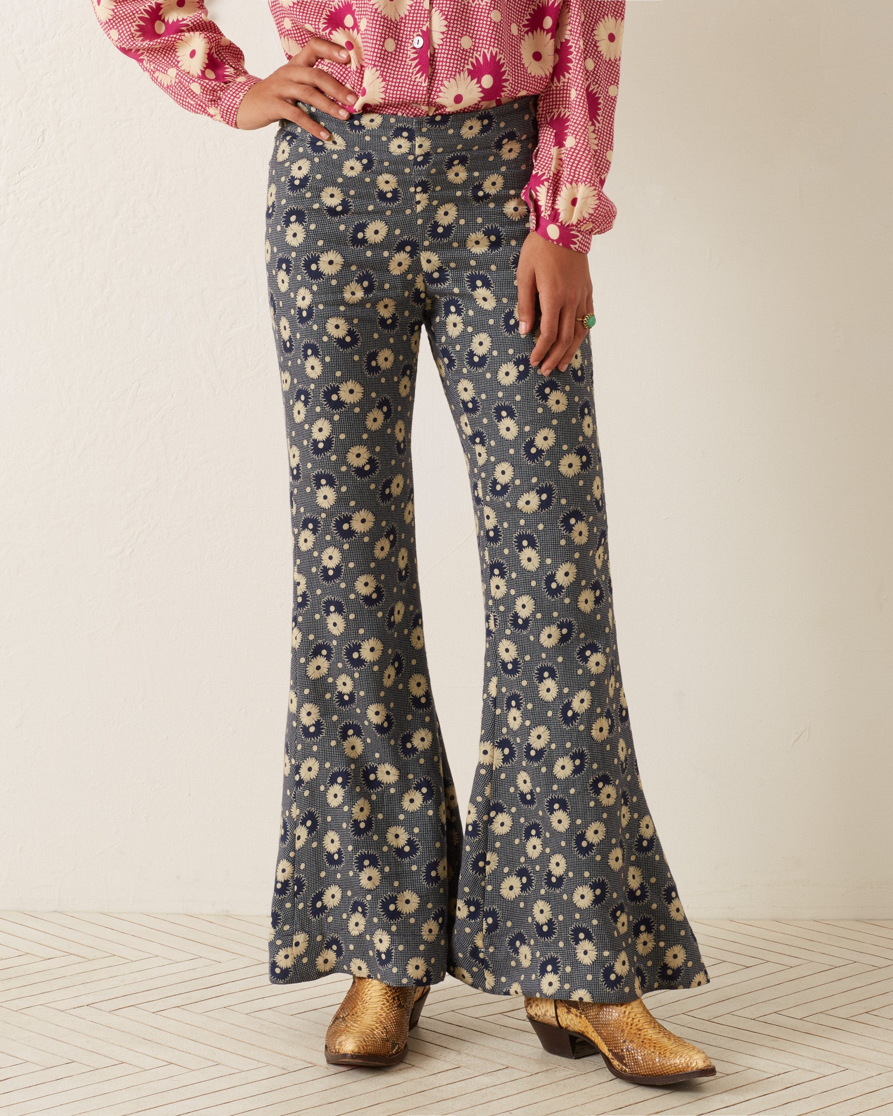 A person wearing the Farah Navy Daisy Pant, featuring high-waisted, '70s-style floral print bell-bottoms, pairs it confidently with a red floral blouse. They complete this retro look with gleaming gold boots, all set against a light-colored floor.