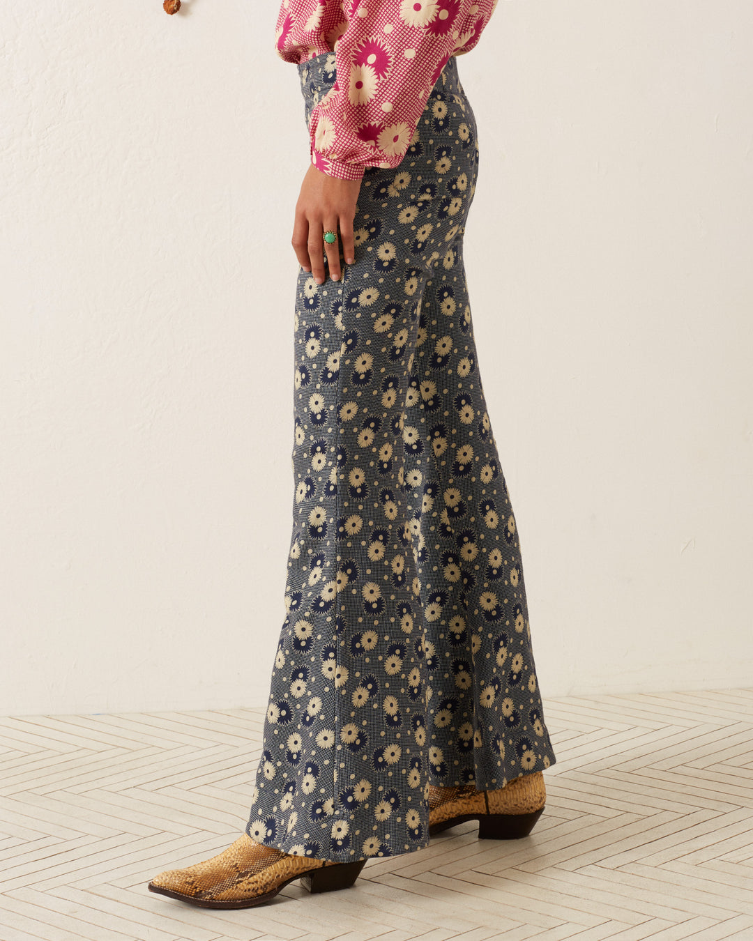 A person wearing Farah Navy Daisy Pants with a '70s-style floral print, a pink top with white flowers, and tan cowboy boots is standing on a light-colored, patterned floor. Only the lower half of the body is visible.