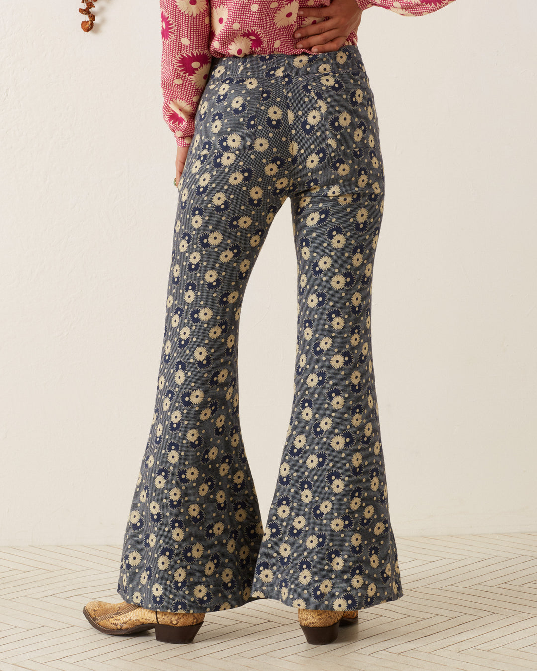 The person is seen from the back wearing the Farah Navy Daisy Pant, a pair of high-waisted, bell-bottom flare pants with a '70s-style floral print. They pair these pants with a pink, long-sleeved top adorned with its own floral design while standing indoors on a light-colored tiled floor.