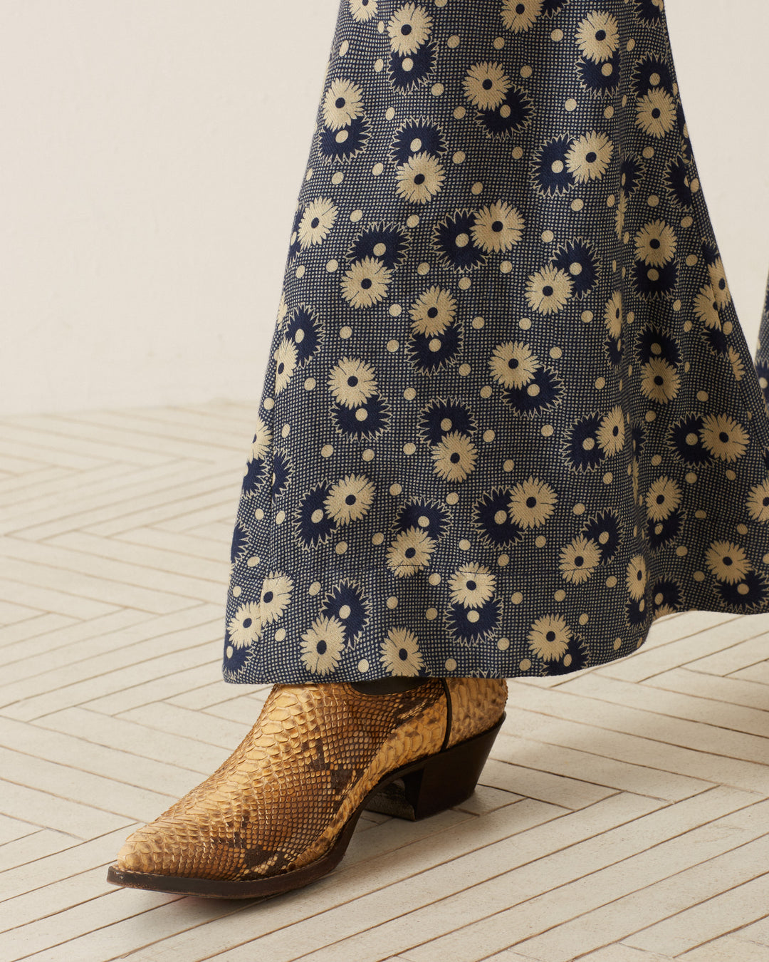 A close-up of a person wearing the Farah Navy Daisy Pant, characterized by '70s-style floral print with a bell-bottom flare in a blue and cream design. They are sporting brown snakeskin boots with pointed toes, standing on a light-colored, herringbone-patterned floor.