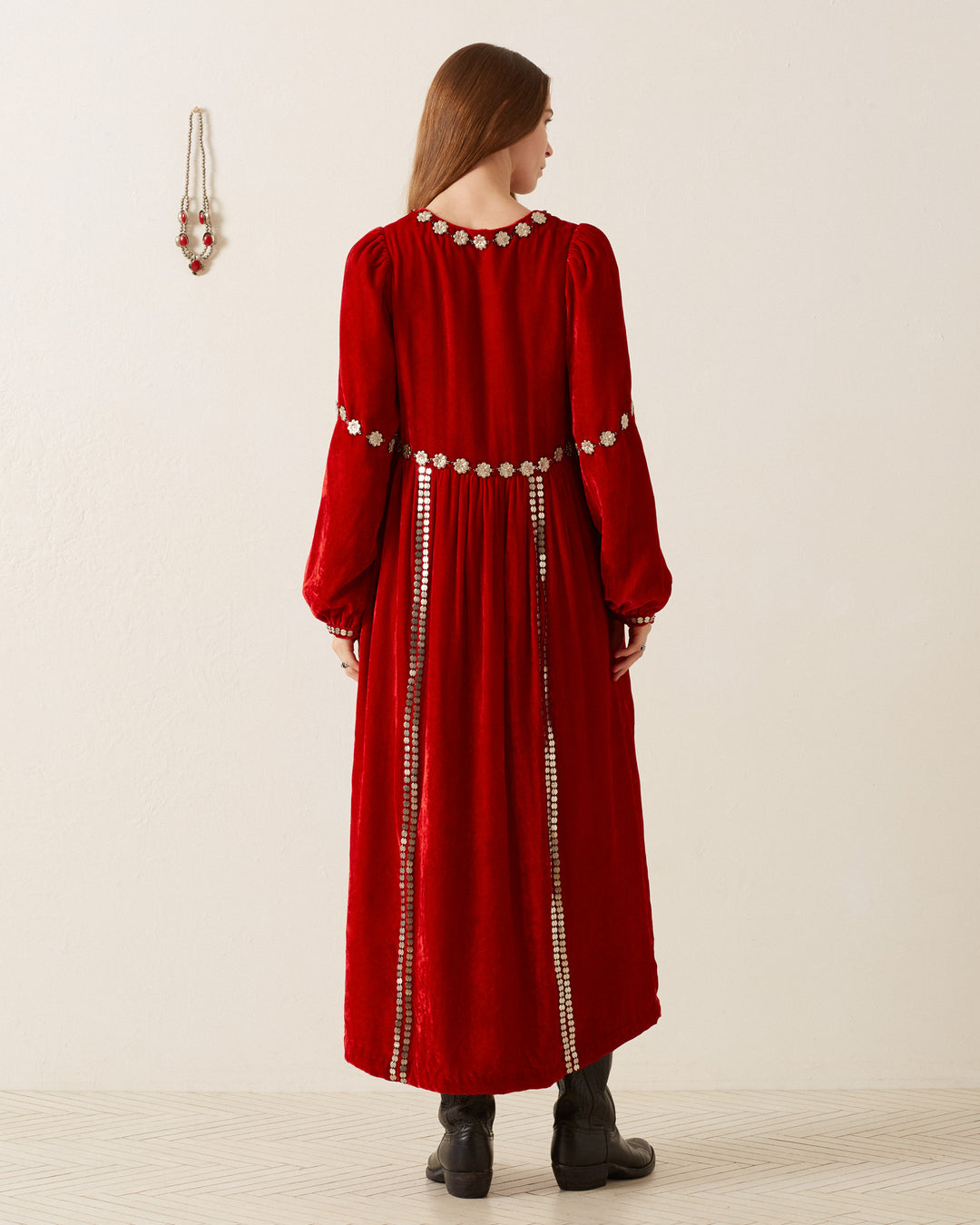 A woman with long hair stands facing away, wearing an Imogen Ruby Velvet Dress, vintage-inspired and adorned with silver circular embellishments. Her black boots complement the look, while on the wall hangs a necklace matching the dress's ornate details.