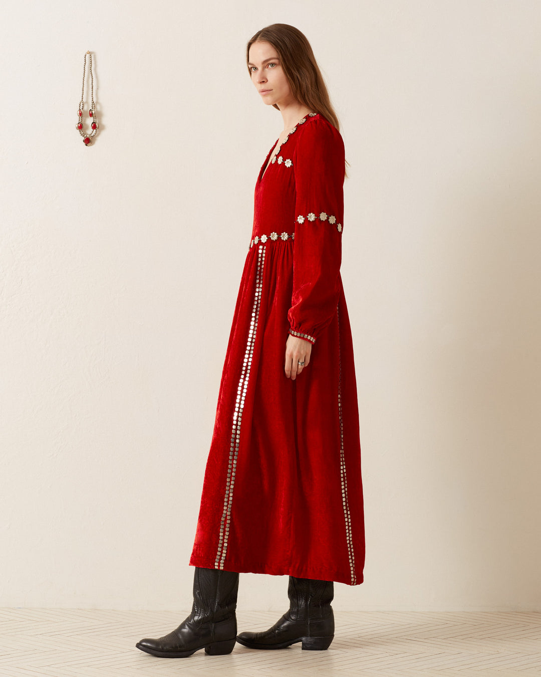 A woman in an Imogen Ruby Velvet Dress with vintage-inspired floral embroidery stands sideways. She is wearing black cowboy boots and has long brown hair. The background is a plain light wall with a small hanging decoration.