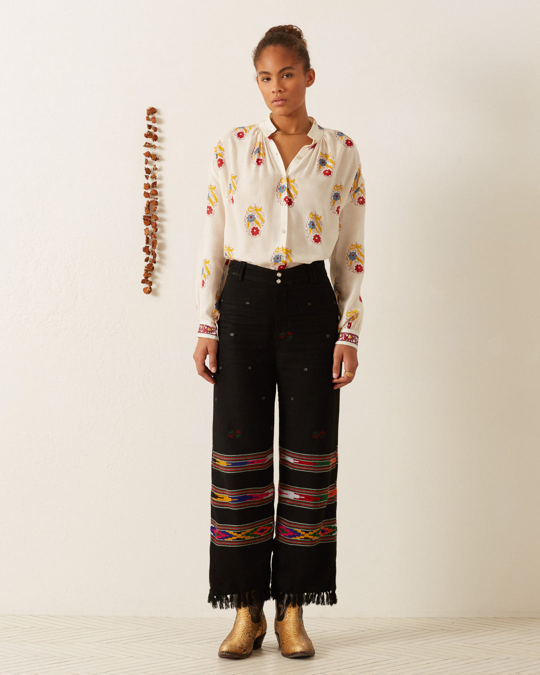 A person stands against a plain background, wearing the Kiki Fireflower Shirt paired with black pants adorned with colorful embroidery near the bottom. Gold boots complete the ensemble. A vertical string of small objects decorates the wall beside them.