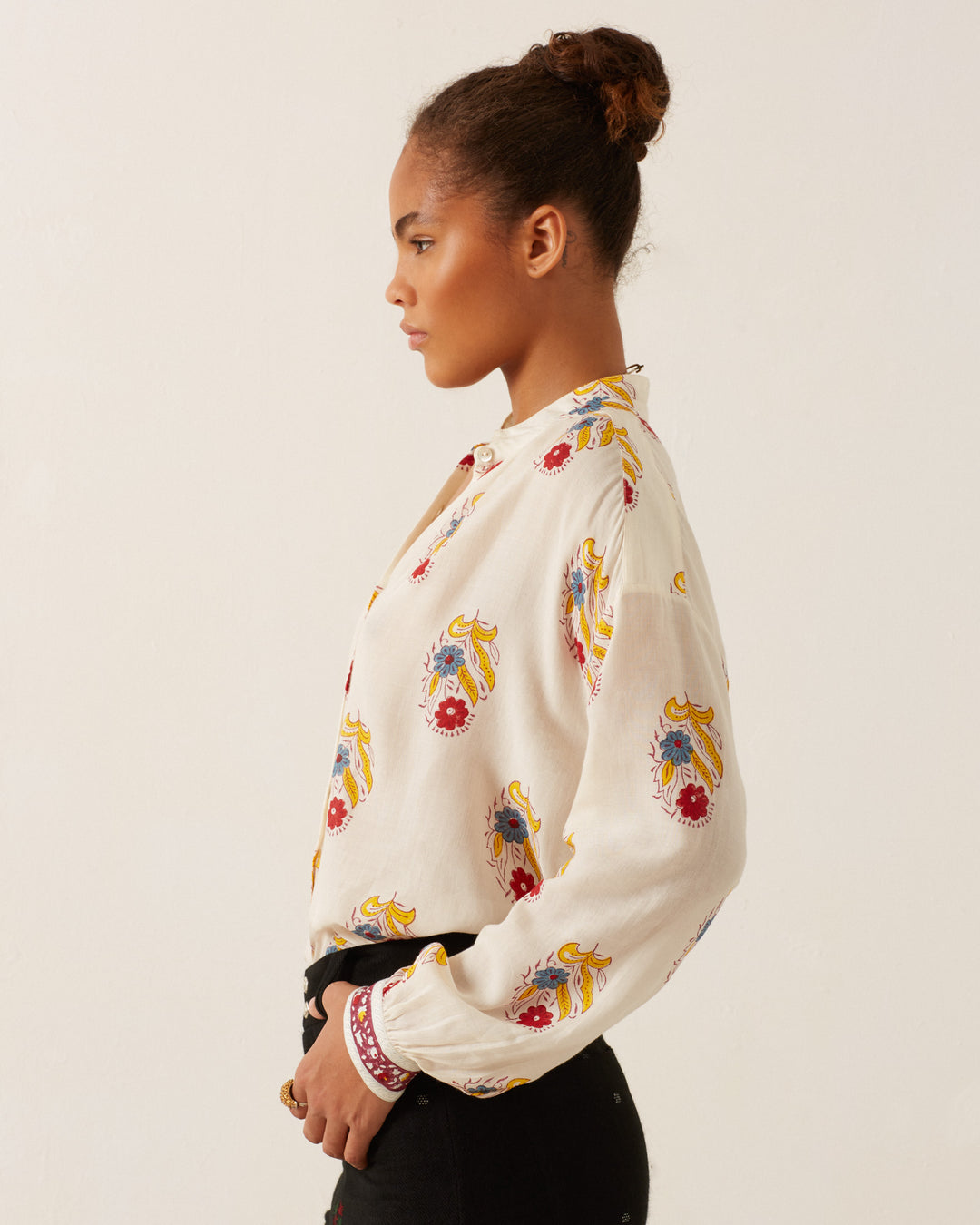 A person with hair in a bun wearing a Kiki Fireflower Shirt stands in profile against a plain background. The shirt, featuring hand-block printed, colorful floral motifs in vibrant red and yellow designs, adds charm to the ensemble. Complementing the shirt, they are also wearing dark pants.