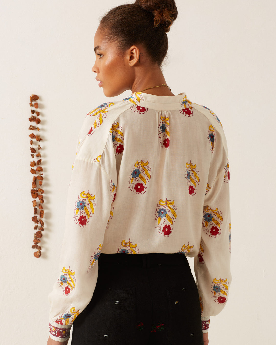 A woman with a bun hairstyle is facing away, wearing the Kiki Fireflower Shirt, which features a hand-block printed paisley pattern in cream. She stands next to a vertical line of dried leaves on the wall. Her ensemble includes dark pants, enhancing the artisanal charm of her attire.