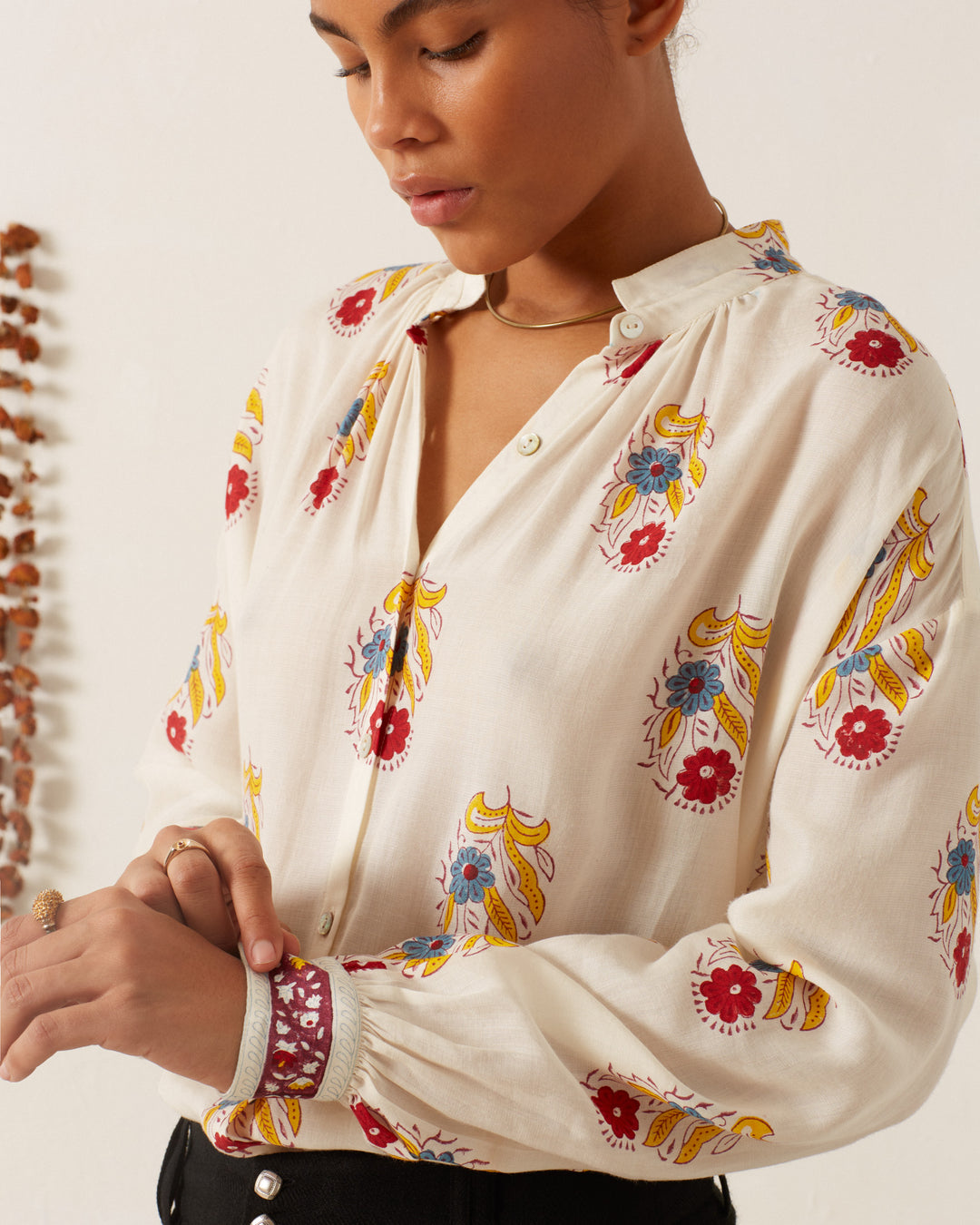 A person wearing the Kiki Fireflower Shirt, a Tencel twill blouse adorned with colorful floral patterns in red, yellow, and blue designs, stands against a light-colored wall while adjusting the cuff of their sleeve.