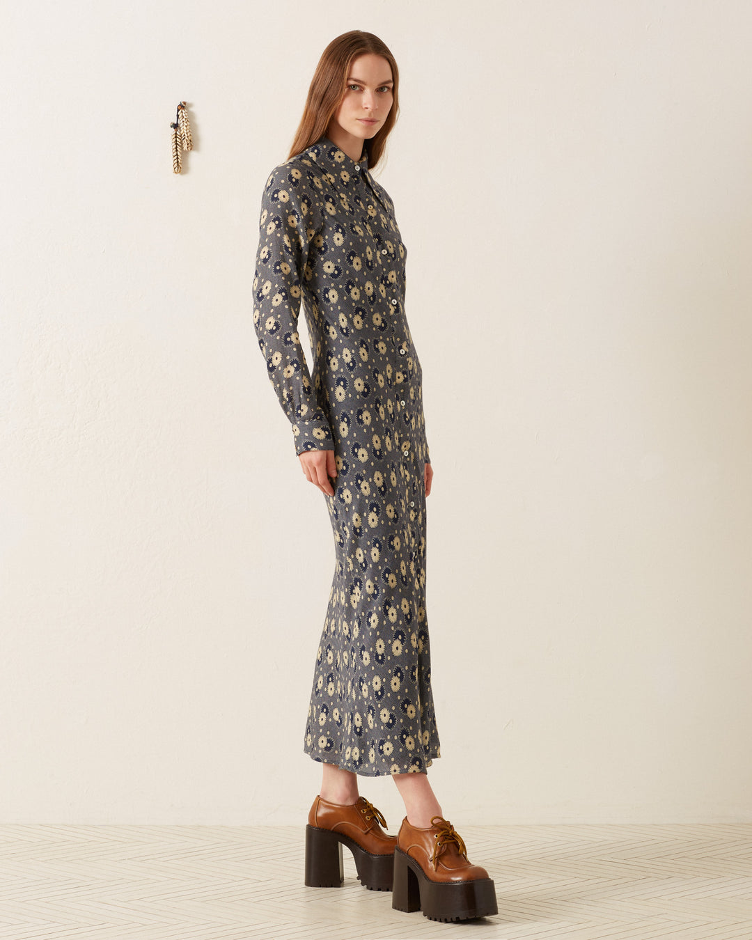 A person with long hair wears the Lotte Navy Daisy Dress, a long-sleeved, ankle-length retro floral shirt dress featuring a '70s-style wing collar, paired with brown platform shoes. They stand against a plain off-white wall adorned with a small decoration, creating a charming vintage ambiance.