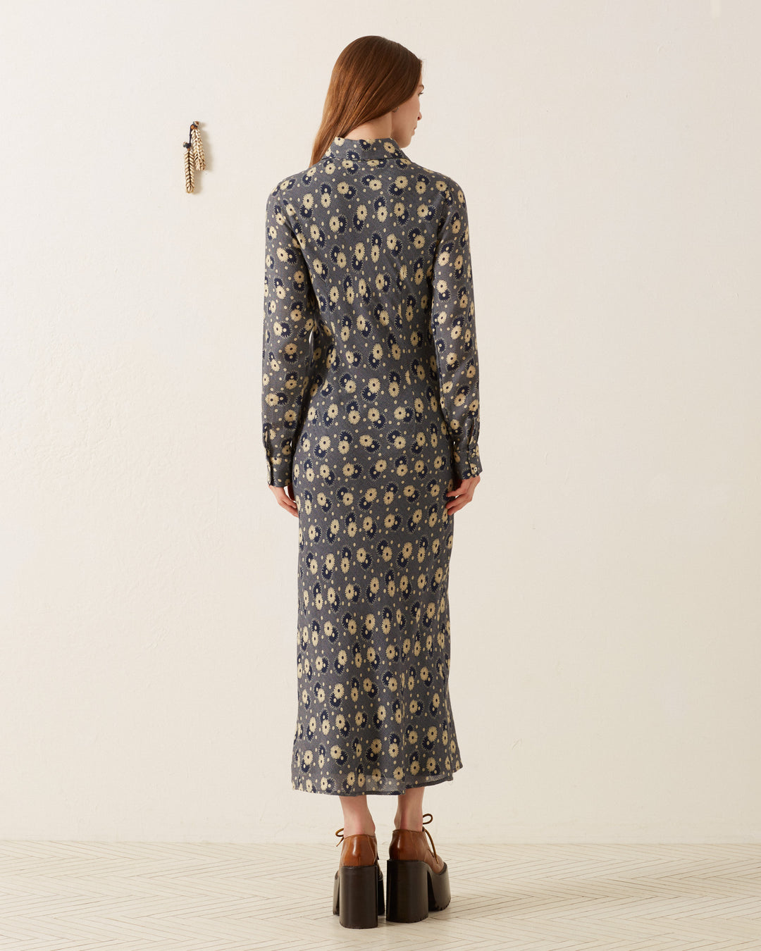 A person with long hair stands facing away, wearing the Lotte Navy Daisy Dress that boasts a '70s-style wing collar and a dark blue and cream retro floral pattern. Set against a light-colored background, they exude a vintage feel, accented by their brown platform shoes.