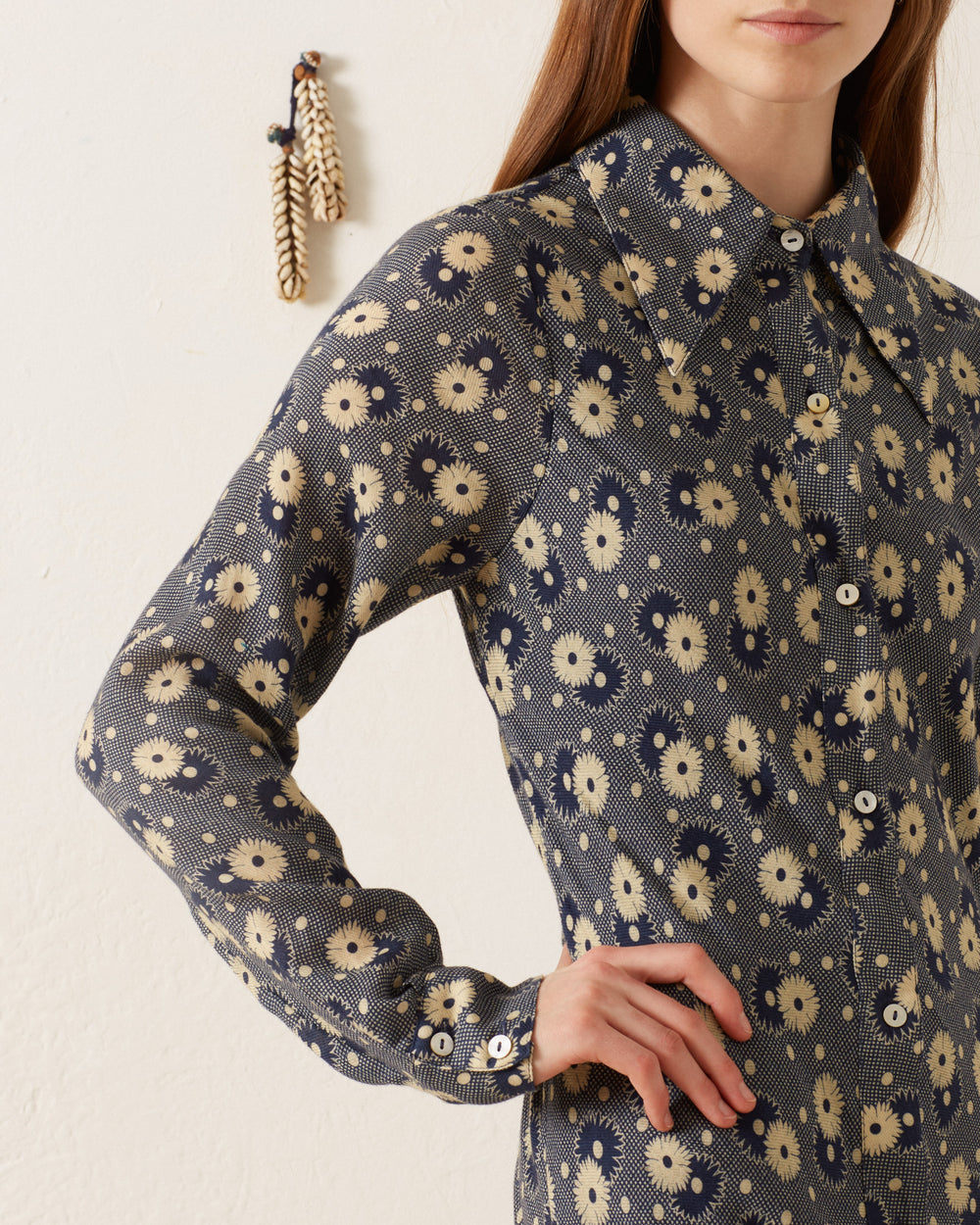 A person is dressed in the Lotte Navy Daisy Dress, a vintage long-sleeved outfit adorned with a navy blue and beige floral pattern. The '70s-style wing collar and buttons enhance its retro charm. In the background, an off-white wall displays an ornament of three tassels, perfectly complementing the nostalgic vibe of the ensemble.