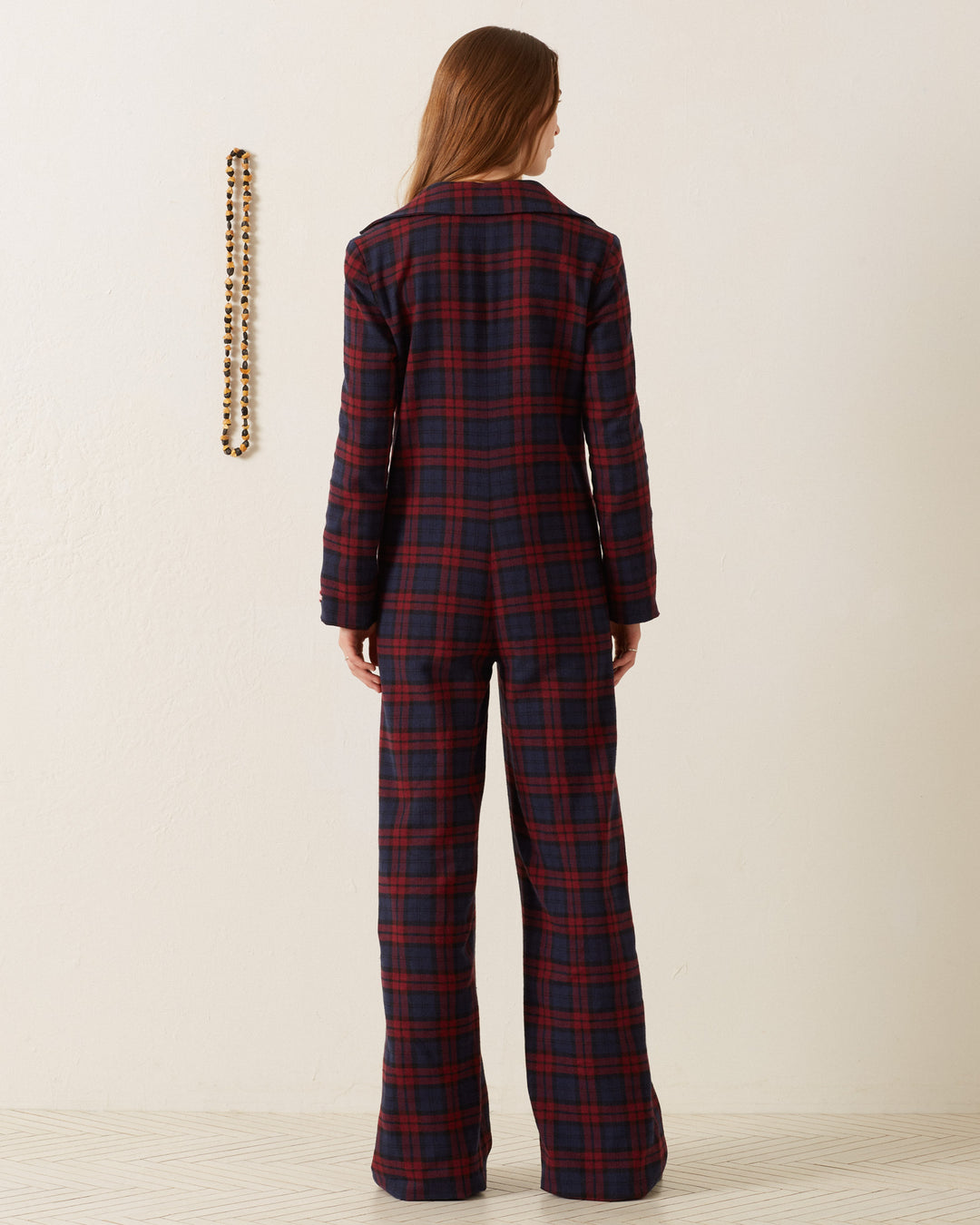 A person with long hair is wearing the Louisa Berry Plaid Jumpsuit, standing with their back to the camera. A necklace hangs on the wall, adding a touch of mystery.