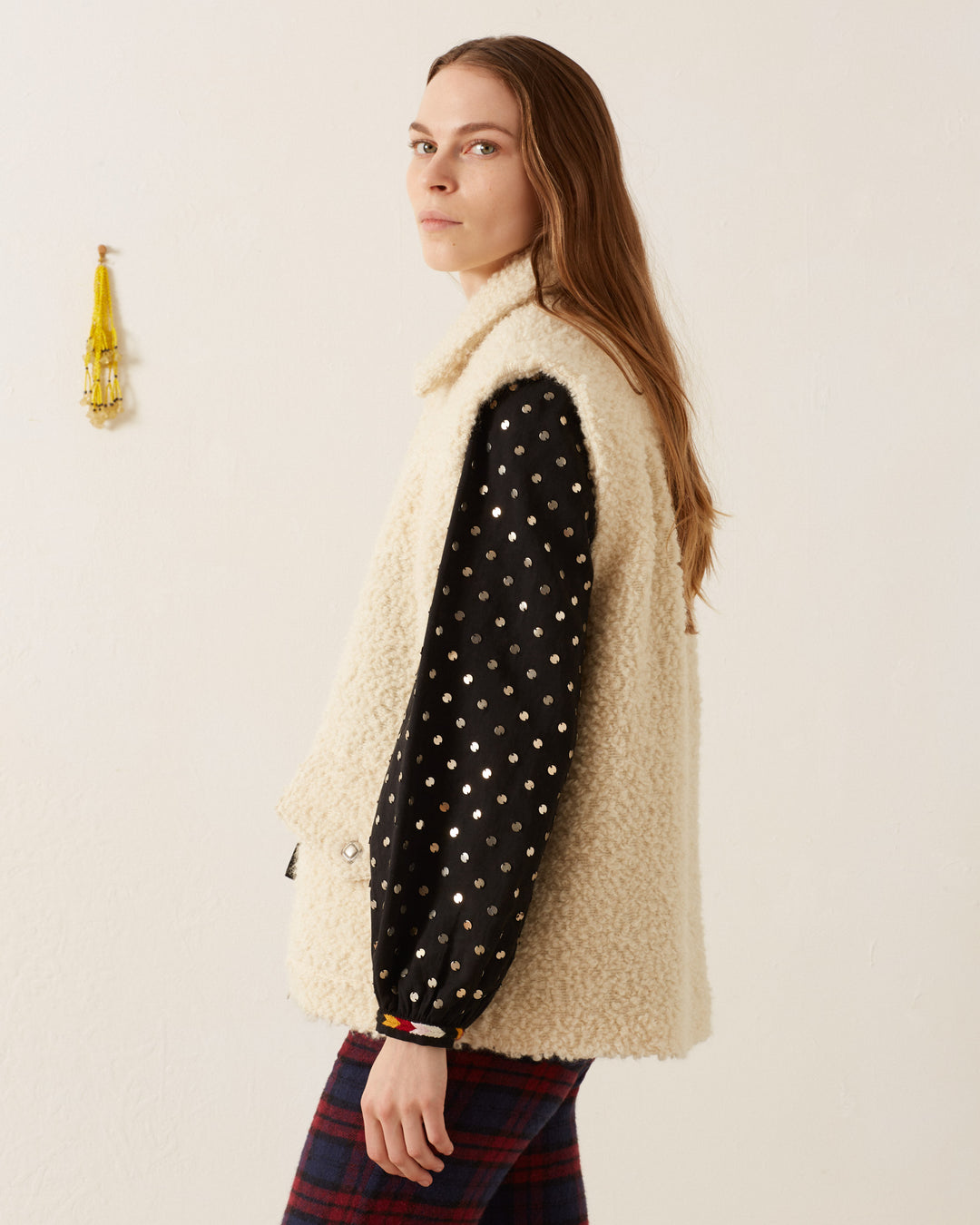 A person with long hair, wearing a polka dot sleeve and the textured Mickey Suri Wool Vest, stands in a minimalist room featuring a small yellow wall hanging.