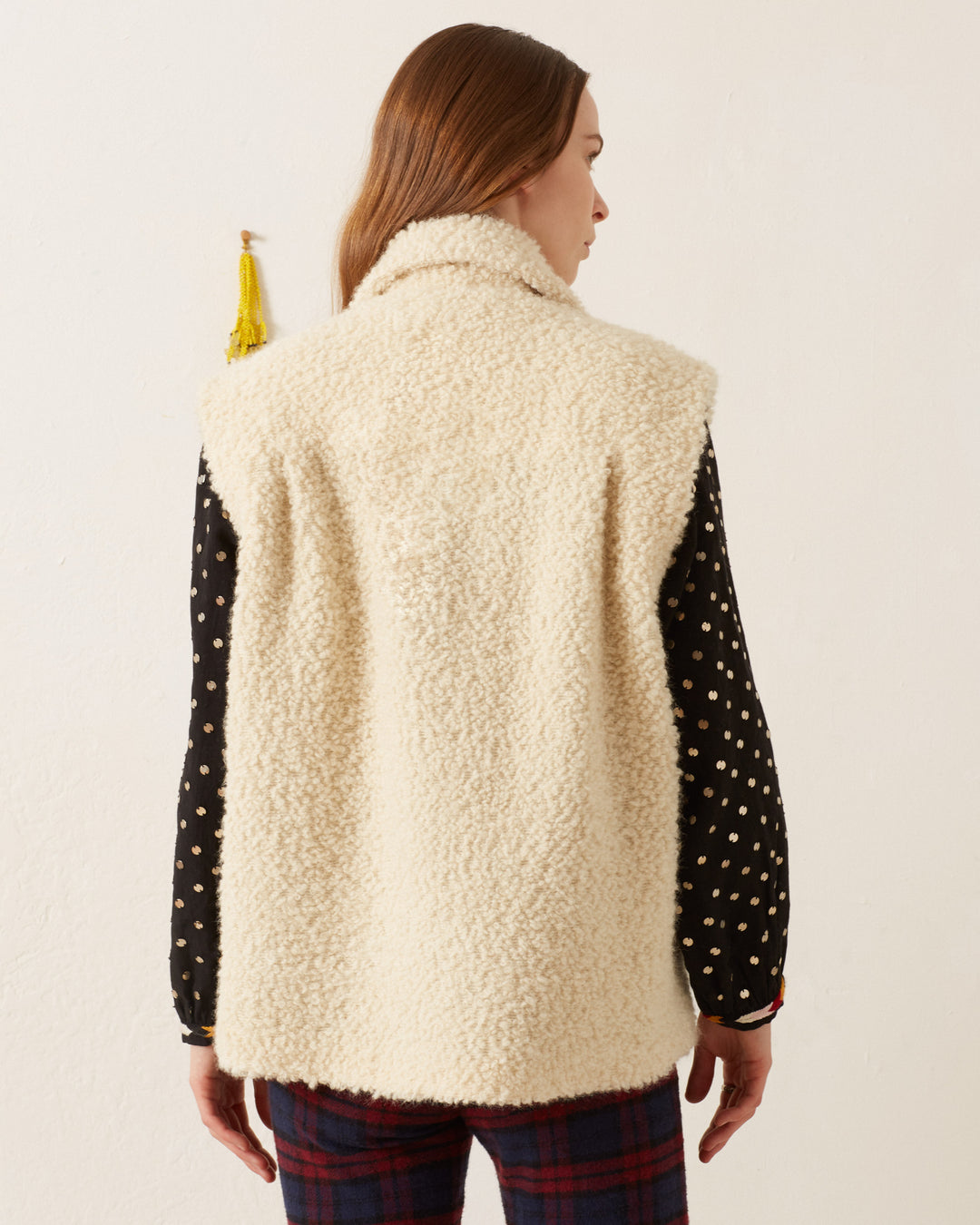 A person is viewed from the back, showcasing the Mickey Suri Wool Vest in a cream color over a black, patterned shirt and plaid pants—perfect for winter layering.