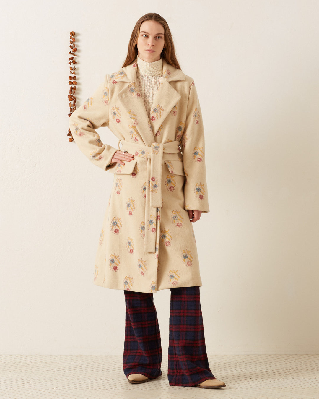 A person stands against a white wall wearing the Officer Fireflower Wool Coat, a long beige wool coat with colorful floral print, paired with a white high-neck top, red and blue plaid pants, and beige shoes. A decorative hanging is visible on the wall.