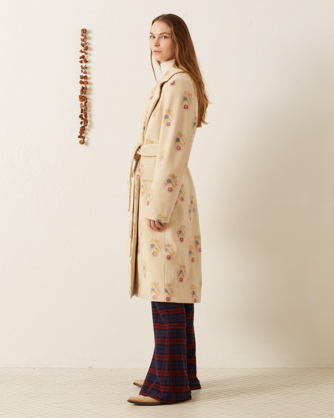 A person with long hair stands sideways, wearing the Officer Fireflower Wool Coat, paired with plaid flared pants and brown shoes. A vertical string of small brown objects hangs on the plain white wall behind them.