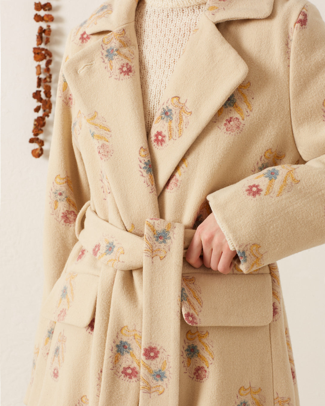 Someone is wearing the Officer Fireflower Wool Coat, a cream-colored wool coat featuring vibrant floral and abstract patterns with a belt. This hand-block printed piece is perfectly layered over a knitted cream sweater, while dried plants hang in the background.