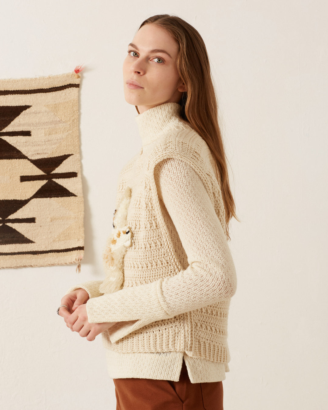 A person with long hair, wearing an Oona Llama Crochet Top in a cream-colored texture, stands next to a wall hanging with geometric patterns, evoking vintage-inspired charm. Against an off-white background, they glance over their shoulder.