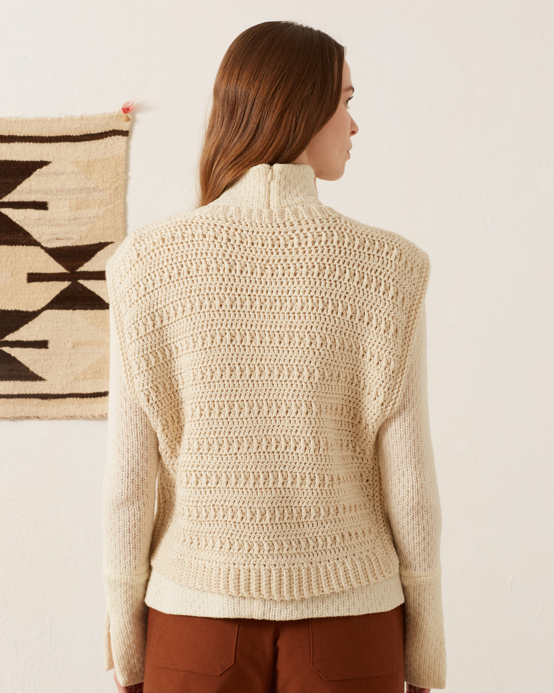 A person with long brown hair stands with their back to the camera, wearing the Oona Llama Crochet Top, a vintage-inspired cream knit sweater showcasing intricate patterns. They are in a room adorned with a beige woven textile hanging on the wall.