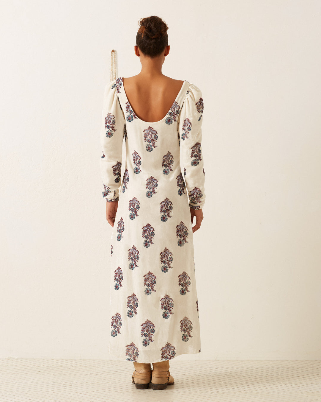 A person with tied-back hair stands with their back to the camera, wearing the Paloma Azure Mirabilis Dress, a long white dress featuring a hand-block printed floral motif. They are positioned against a plain off-white background.