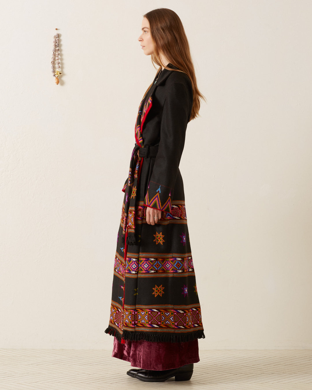 A woman with long brown hair stands in profile, wearing the Penelope Wildflower Wool Coat—a handcrafted garment featuring colorful geometric designs and a belted waist. She pairs it with black shoes and a maroon skirt underneath, while a decorative object hangs on the wall.