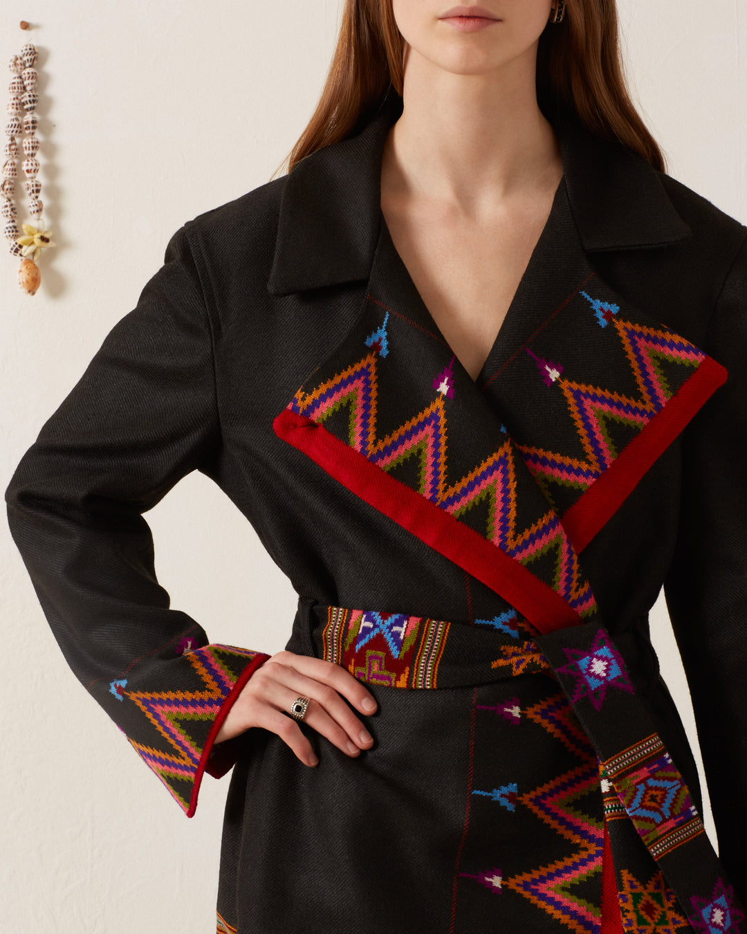 A person wearing the Penelope Wildflower Wool Coat, which features colorful geometric patterns and a red trim, stands against a light background. They pose confidently, one hand on their hip adorned with a ring. The handcrafted garment adds charm to the scene, complemented by an ornamental decoration hanging on the wall.