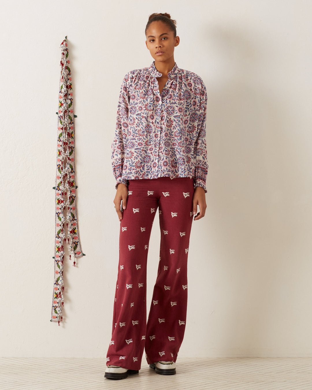 A person stands against a plain wall, dressed in red pants with small designs and wearing a Poet Amazon Floral Blouse. A floral scarf hangs on the wall beside them, complementing the light wood floor.