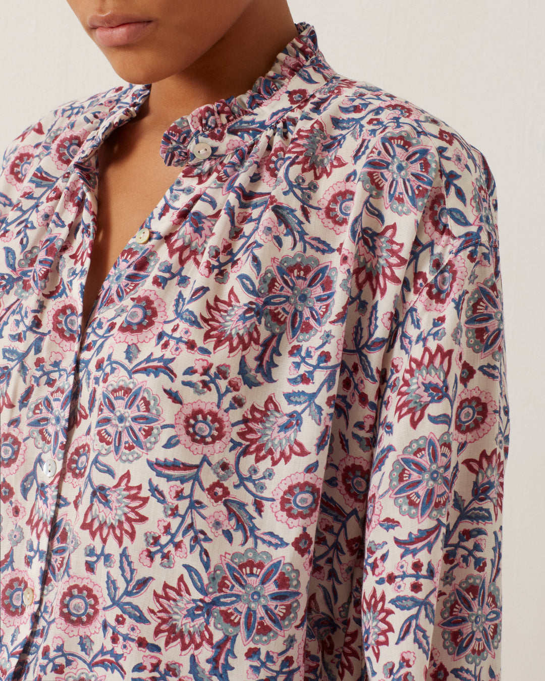 A person dressed in the Poet Amazon Floral Blouse, adorned with vintage-inspired red, blue, and white flowers, is visible. The blouse boasts a ruffled collar and buttons down the front. The person's face is partially visible against a plain, light background.