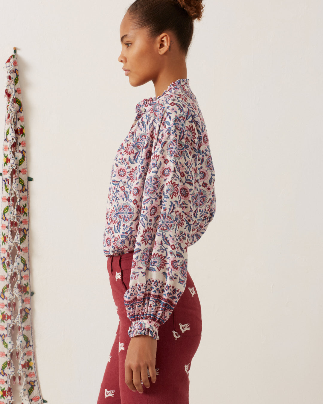 A person with a bun hairstyle stands sideways, wearing a Poet Amazon Floral Blouse in Tencel twill and maroon pants adorned with white designs. A patterned cloth hangs on the left side against a plain wall.