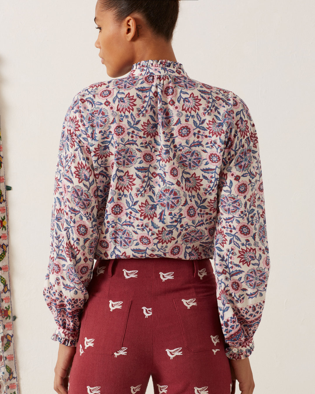 A person stands facing away, wearing the Poet Amazon Floral Blouse, a vintage-inspired piece with ruffled cuffs and collar, paired with maroon pants adorned with bird designs. The background is a light-colored wall that showcases another floral fabric.