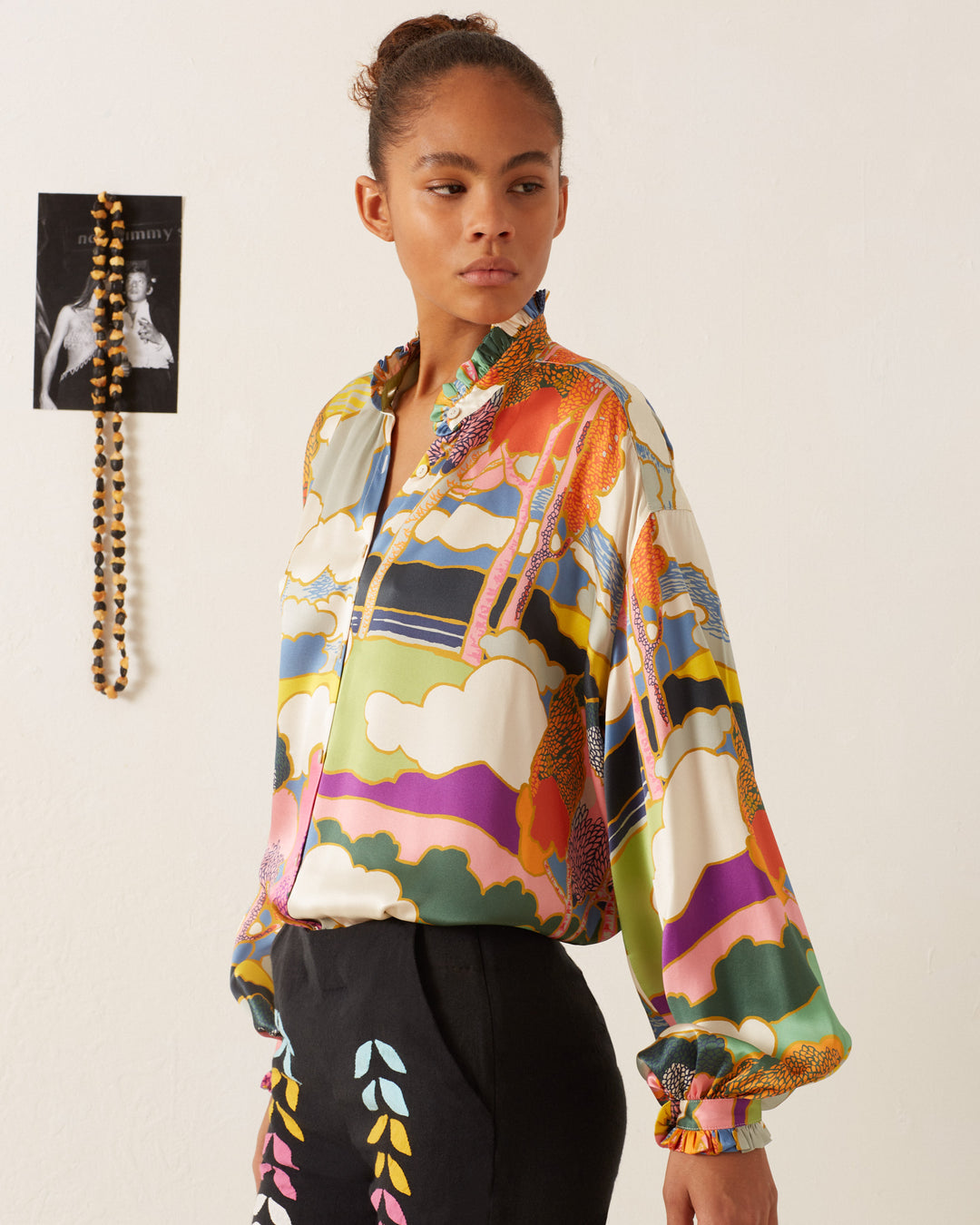 A person wearing a vibrant Poet Aurora Silk Blouse and black pants stands in a room where an Art Nouveau landscape print graces the wall, alongside a beaded necklace.