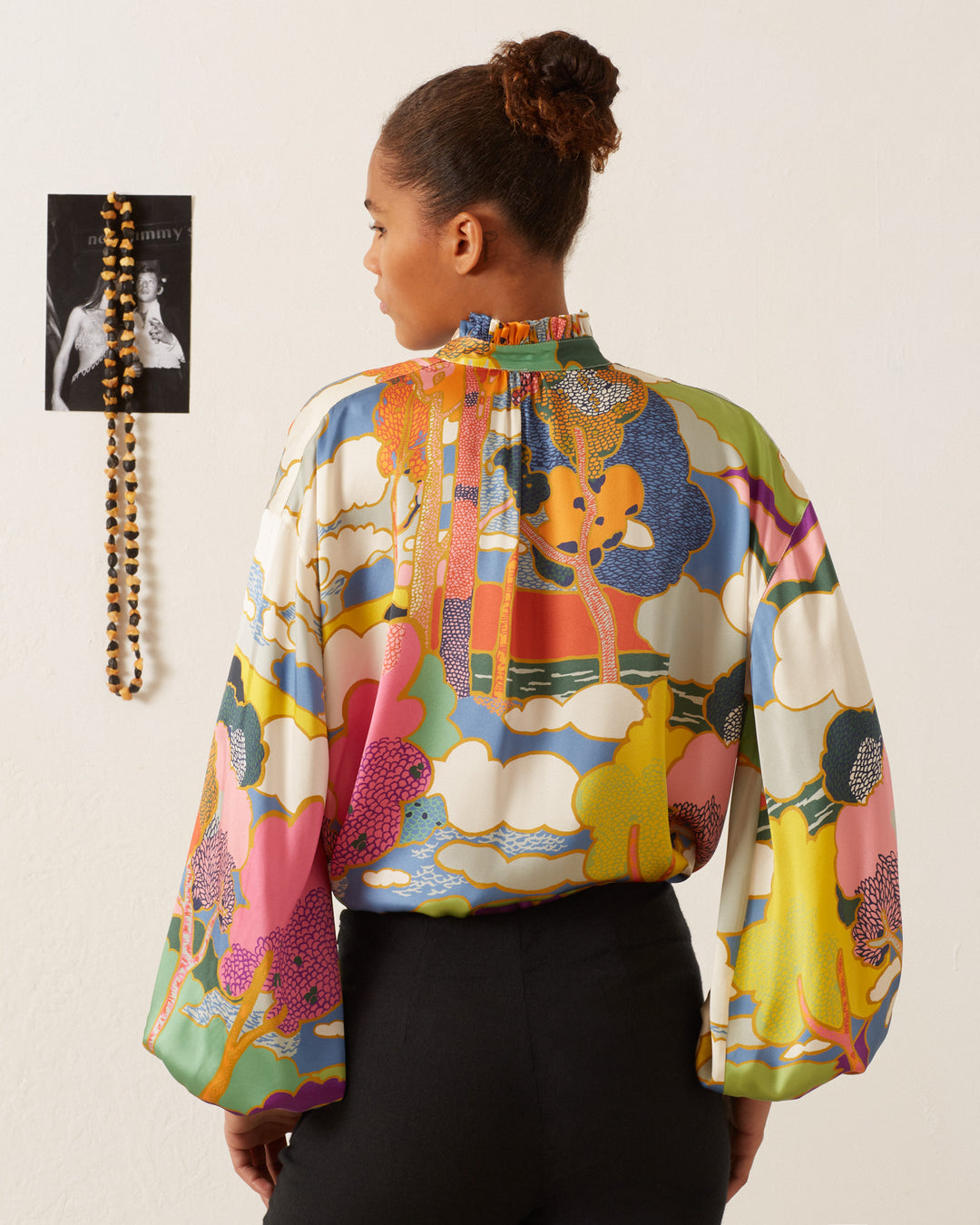 A person wearing a Poet Aurora Silk Blouse with billowy sleeves stands facing away. A photo and beaded necklace hang on the wall, complementing the room's Art Nouveau landscape print.