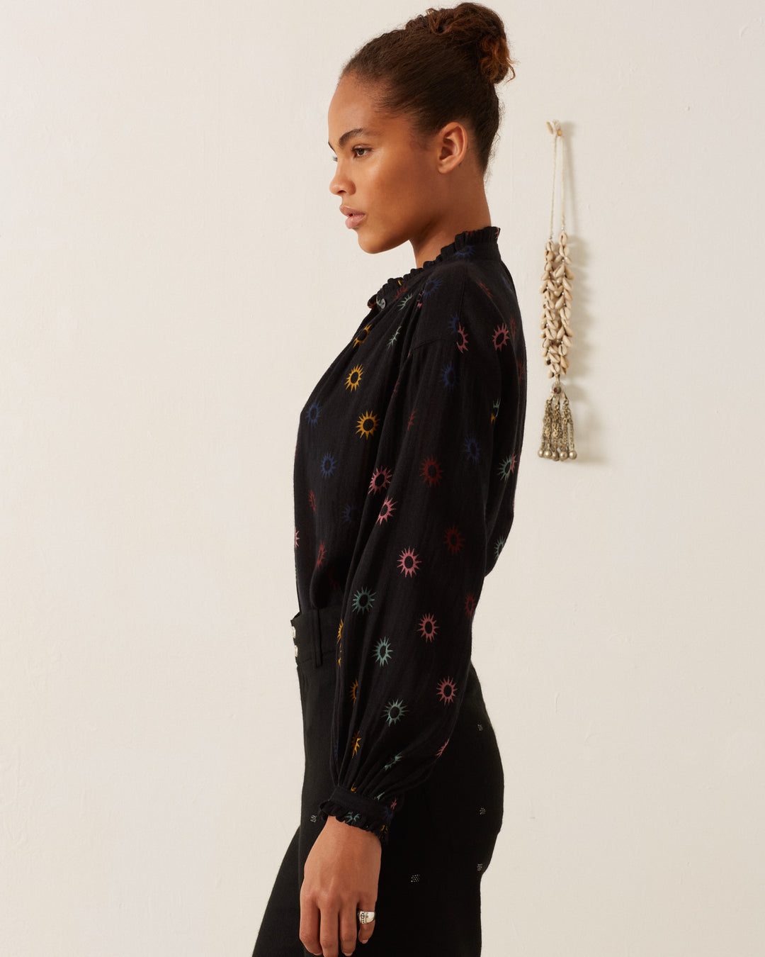 A woman in a Poet Evening Star Blouse stands sideways against a light wall with a hanging decoration.