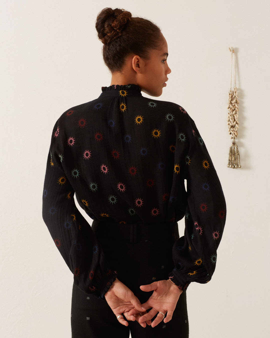 A person with their hair in a bun, wearing the Poet Evening Star Blouse featuring vibrant celestial prints, stands facing away from the camera against a plain wall with a decorative tassel hanging.