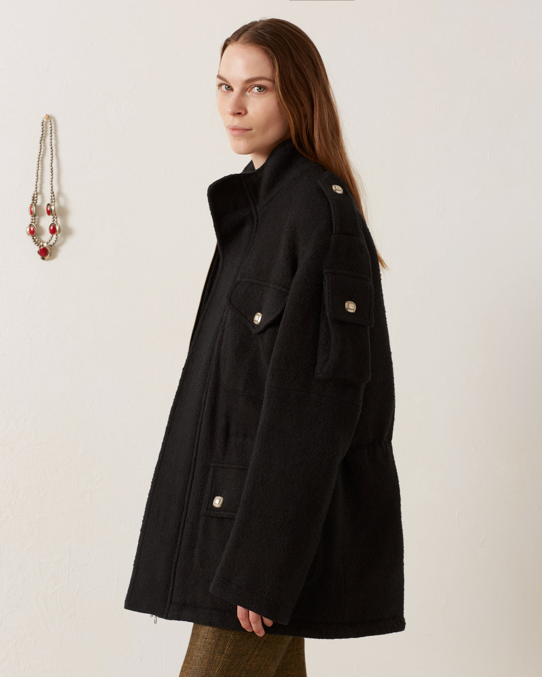 A person with long brown hair is wearing a Porter Sable Llama Coat, characterized by its black color and buttoned pockets, while standing against a white wall. To the left, a necklace adorned with red and silver beads hangs on the wall.