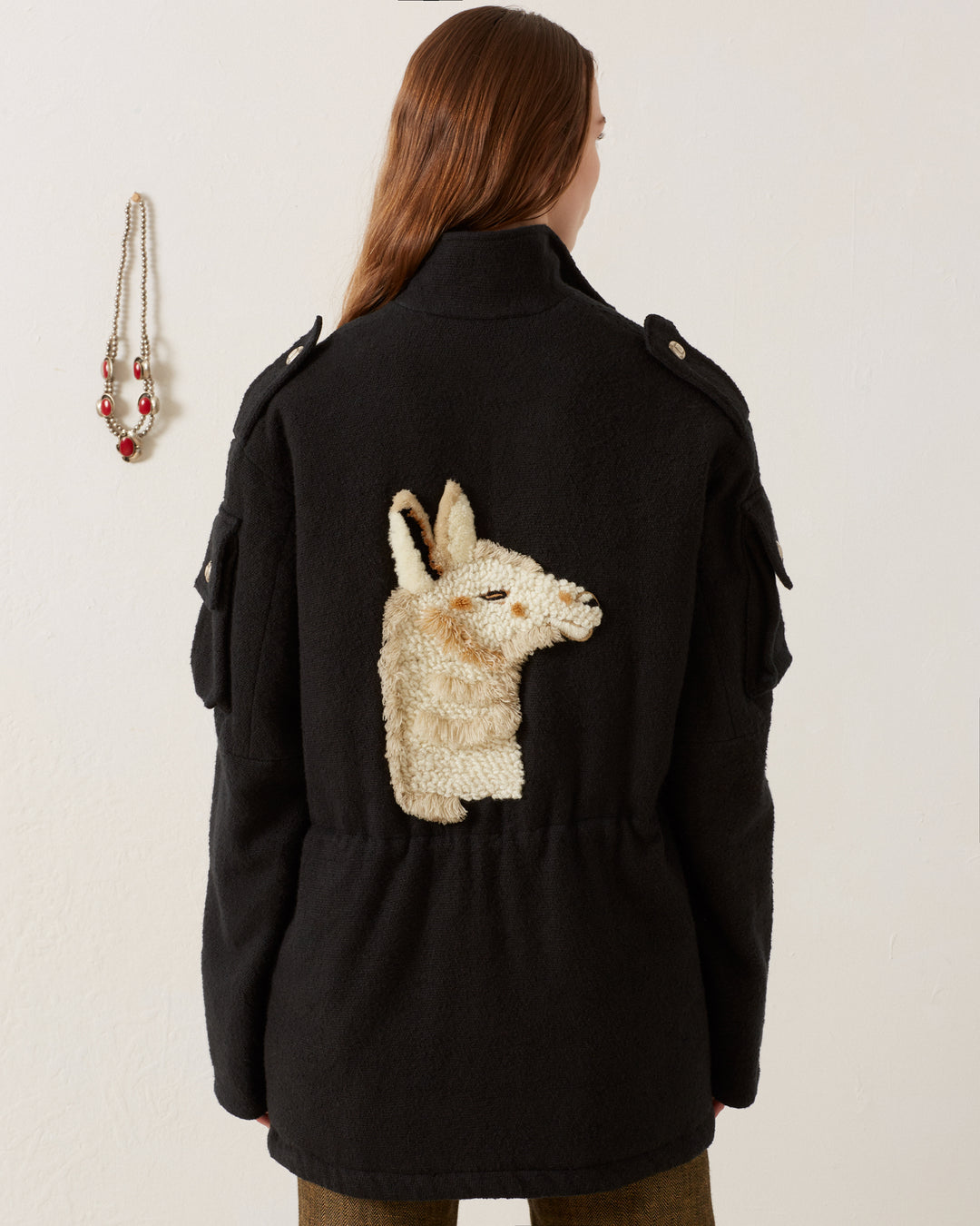 A person with long hair wears the Porter Sable Llama Coat, a black utility jacket made of Khadi cotton with epaulets and side pockets, featuring a textured animal head design on the back. A beaded necklace with red accents hangs on the wall.