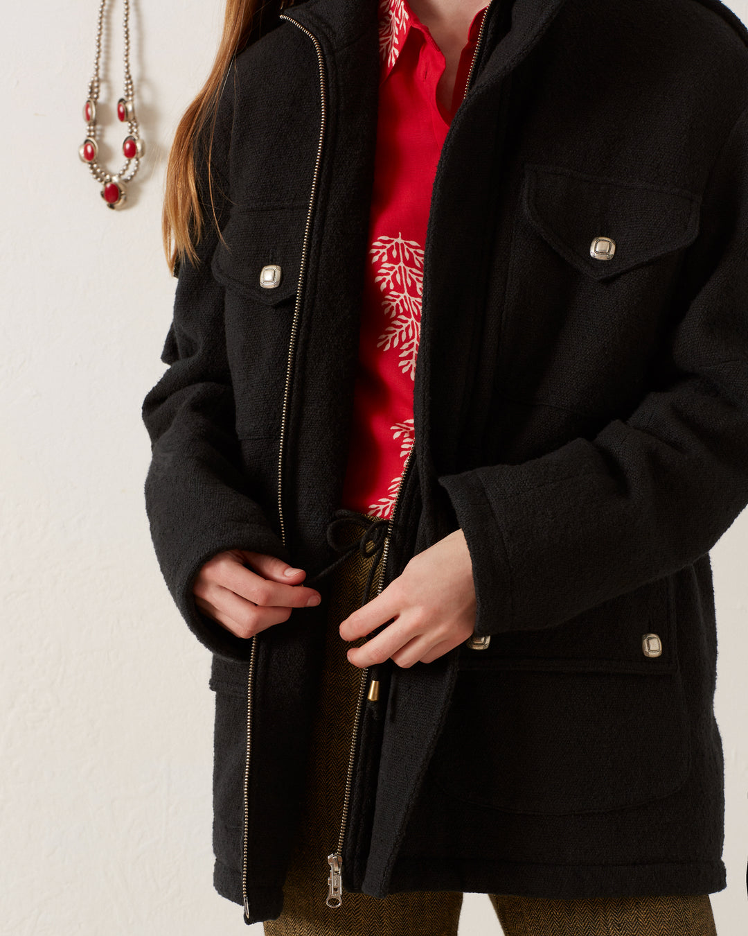 A person wearing a Porter Sable Llama Coat over a red shirt with white patterns zips up their coat. They are also wearing brown pants. A necklace with red beads hangs on a hook on a white wall in the background, adding to the scene's charm.