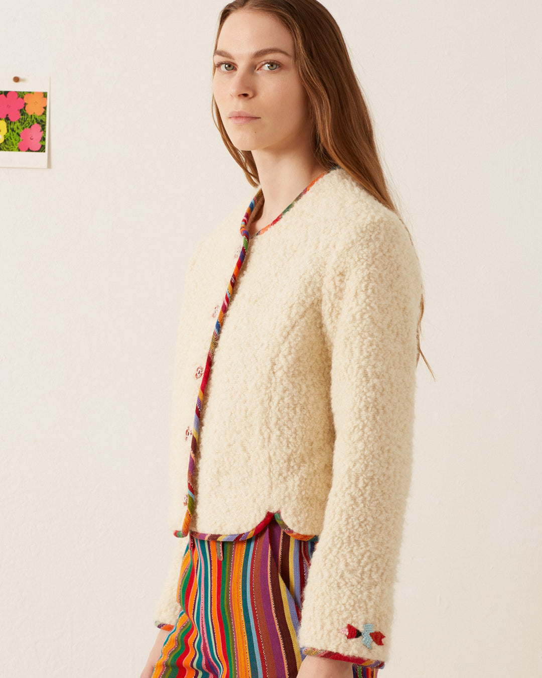 A person with long brown hair is wearing the Rowan Suri Wool Jacket, crafted from cream alpaca wool, featuring embroidered details and handmade floral buttons. The jacket boasts a scalloped hem and is paired with colorful striped pants. A small floral artwork can be seen on the white wall behind them.