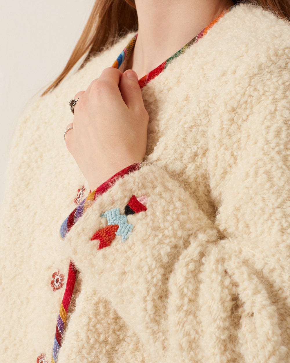 Close-up of a person wearing the Rowan Suri Wool Jacket, featuring a fluffy, cream-colored alpaca-wool design with multicolored embroidered patterns. The collar showcases a vibrant Guatemalan ikat trim, while the sleeve is embellished with handmade floral buttons.