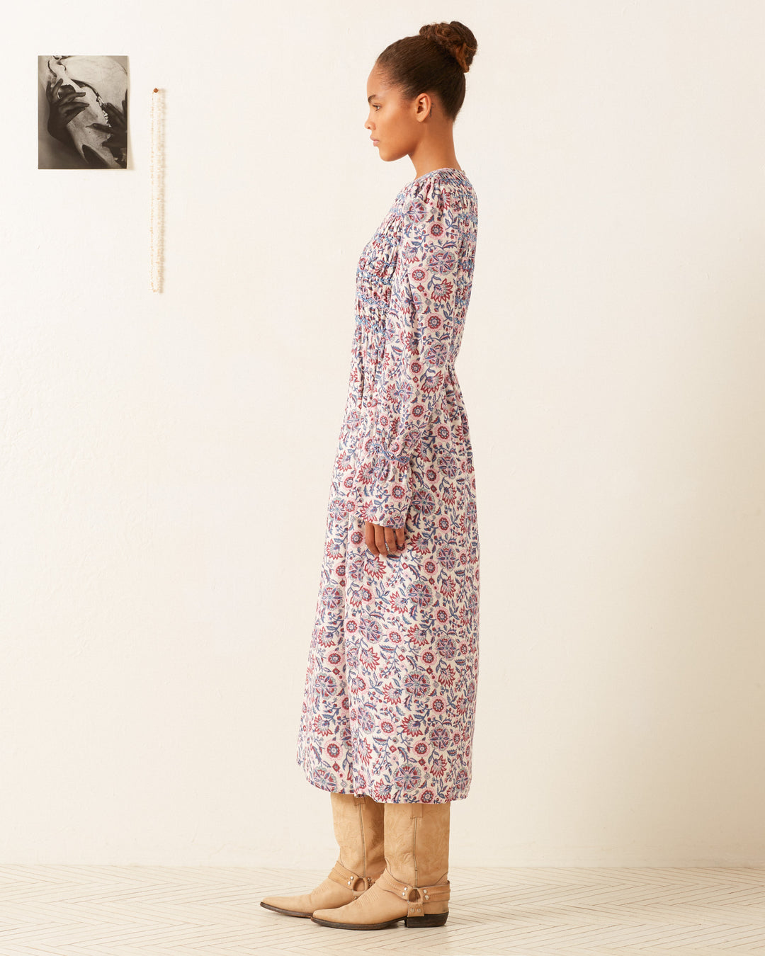 A person standing in profile against a neutral background, wearing the Sabine Amazon Floral Dress paired with tan knee-high boots. There is a small black-and-white photo on the wall.