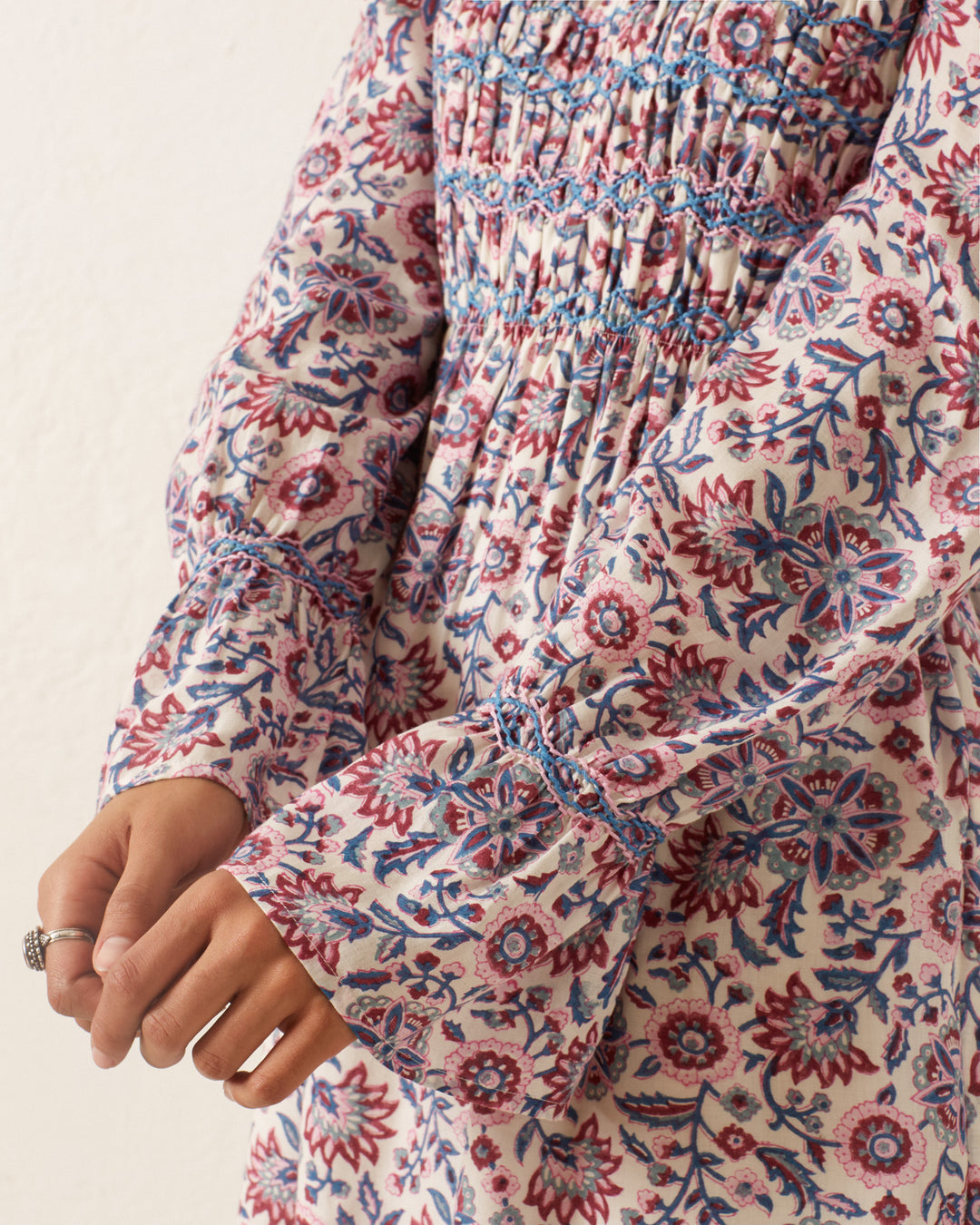 The individual is dressed in the Sabine Amazon Floral Dress, a vintage-inspired garment with long sleeves that showcase red and blue flowers against a white background. A closer look highlights the hand-smocked bodice and a ring on one hand, exuding timeless elegance.