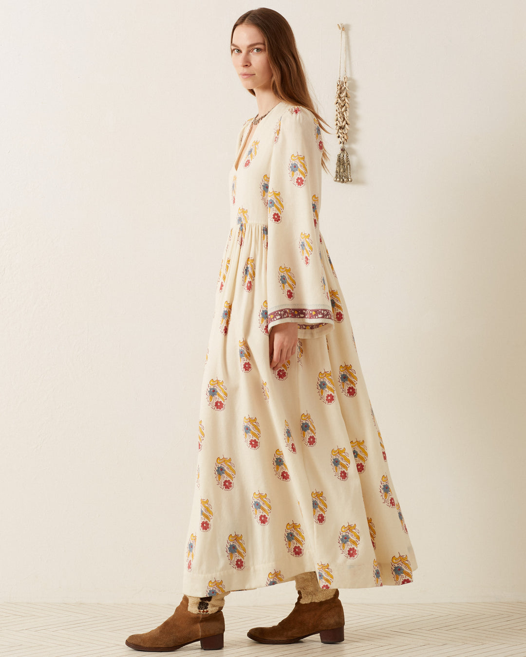 A woman with long hair stands in a room wearing the Savannah Fireflower Wool Dress, which is cream-colored and hand-block printed with colorful patterns and features bell sleeves, paired with brown ankle boots. A tassel decoration hangs on the white wall behind her.