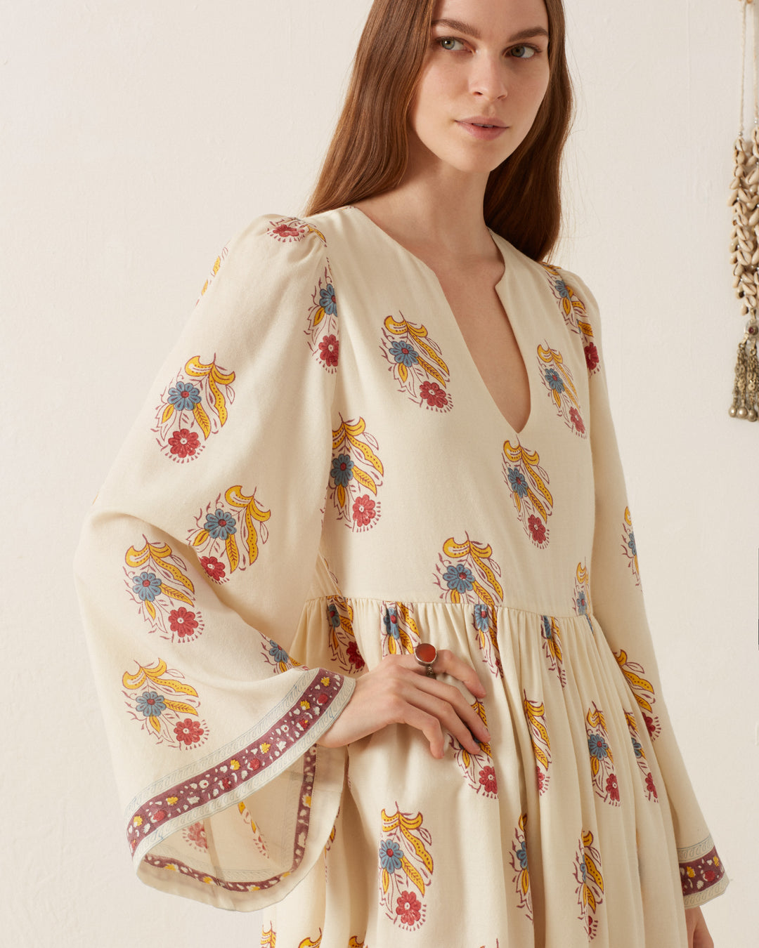 A woman is wearing the Savannah Fireflower Wool Dress, which features a hand-block printed floral motif, long sleeves, and a V-neckline. She stands against a light background, gazing off to the side.