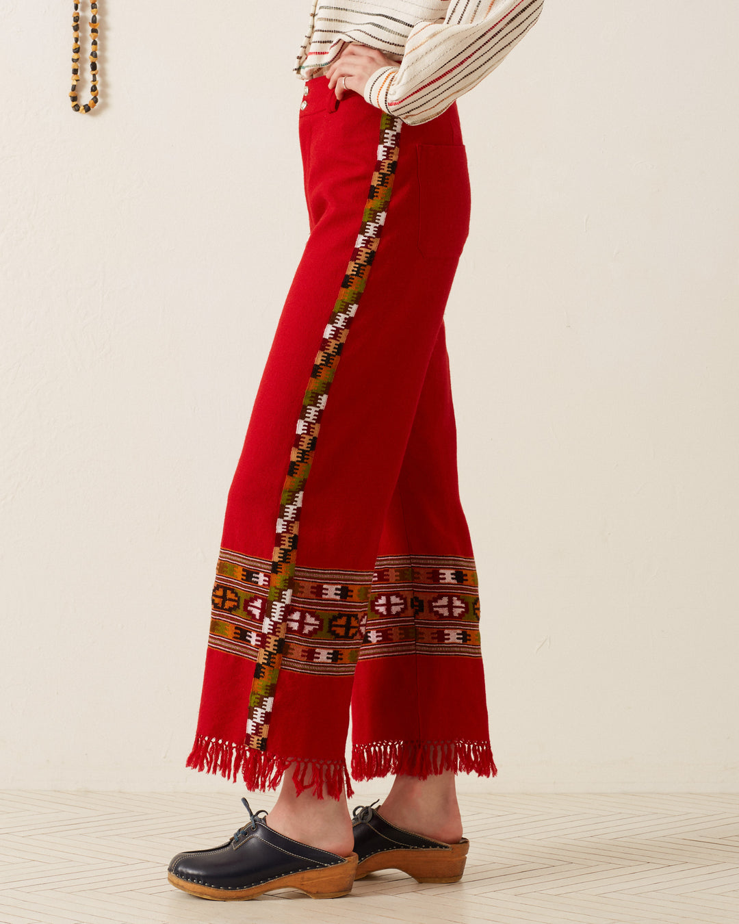 A person wearing the Scout Scarlet Blanket Pant, featuring colorful tribal patterns inspired by Himalayan weaving tradition along the sides and cuffs, paired with black clogs. These pants are crafted from hand-loomed wool and include a fringed hem. The individual is standing sideways against a neutral background.