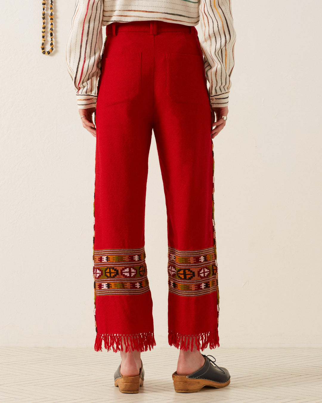 A person wearing the Scout Scarlet Blanket Pant, featuring a bright red color with a fringed hem and vibrant embroidered designs inspired by Himalayan weaving traditions, paired with a striped sweater and brown clogs, is standing indoors against a neutral-colored wall and light flooring.