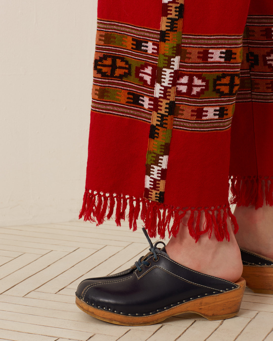 Close-up of a person wearing a pair of black clogs with wooden soles, standing on a light-colored tiled floor. They are dressed in the Scout Scarlet Blanket Pant, showcasing intricate multicolored embroidery inspired by the vibrant Himalayan weaving tradition.