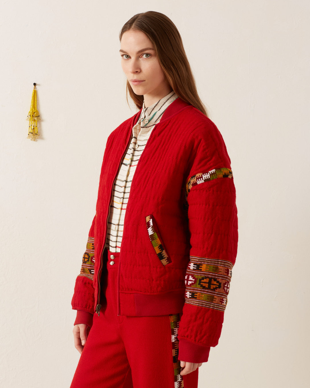 A person wearing the Sutton Scarlet Blanket Bomber, a hand-quilted red jacket with patterned trim, over a striped shirt and matching red pants stands against a plain wall with a small hanging decoration.